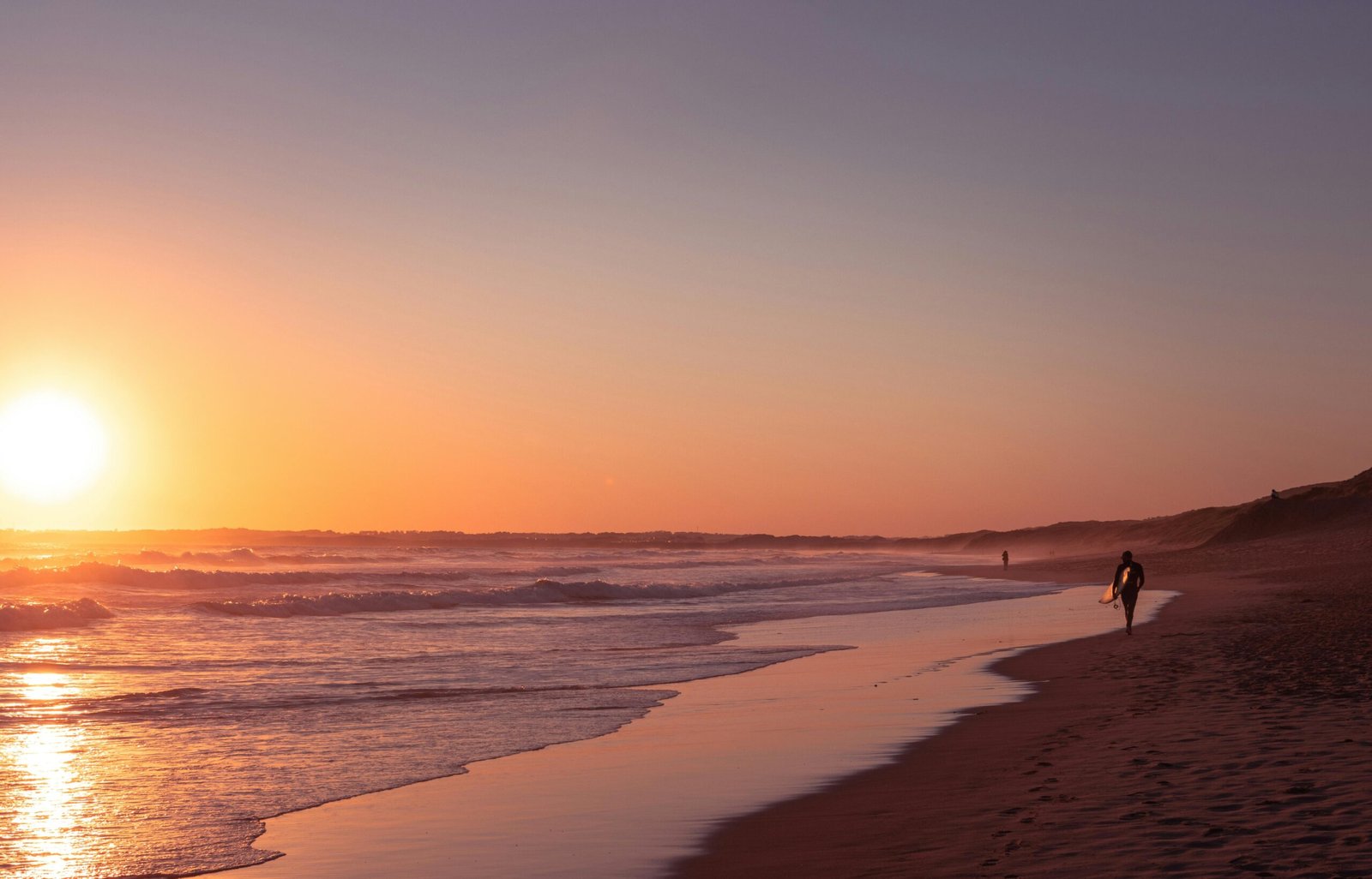 A lone surfer walks along a serene Australian beach at sunset, capturing a peaceful moment by the sea.